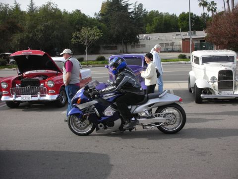 1998 Ford Crown Victoria - helmeted rider on a purple Suzuki Hayabusa motorcycle rolls past a red classic coupe and white hot rod at a car meet.