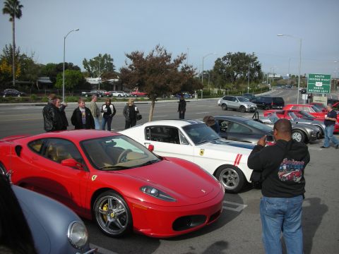 1998 Ford Crown Victoria - red Ferrari coupe sits in a parking lot among spectators and other sports cars near a freeway sign.