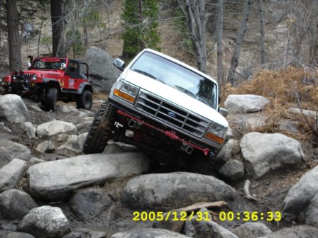 1996 Ford Bronco - Two off-road vehicles tackle a rocky trail in a wooded area.