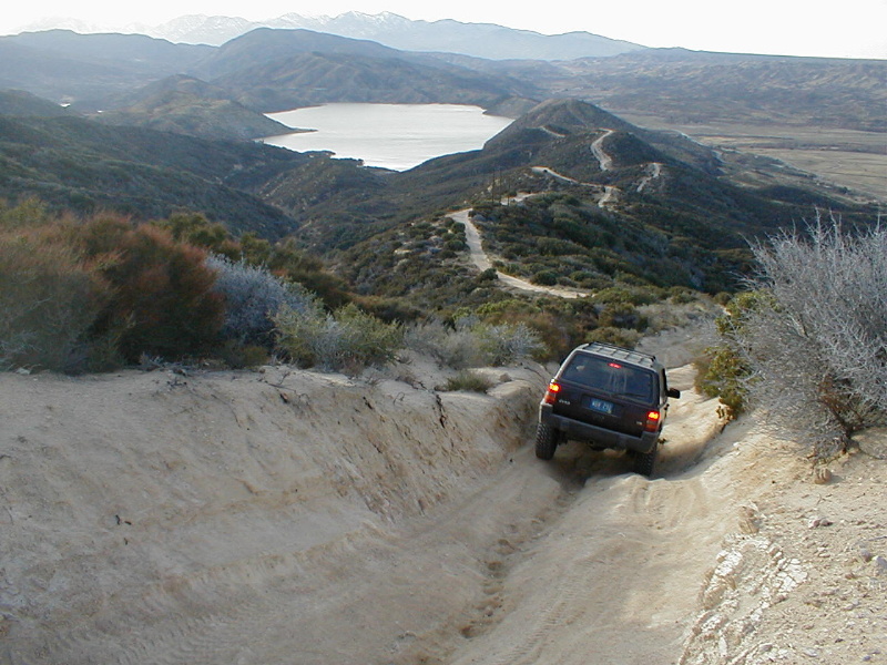 Jeep Grand Cherokee driving down a steep dirt trail with a lake and mountains in the distance