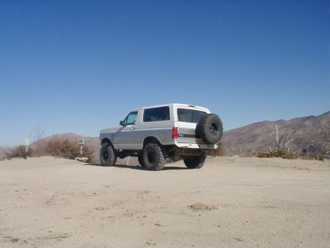 1996 Ford Bronco - A lifted Ford Bronco sits on a dusty trail under a clear blue sky, with mountains in the distance.