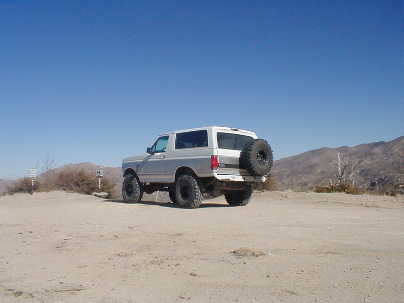 Silver Ford Bronco with spare tire on back, parked on dirt road with hills in background