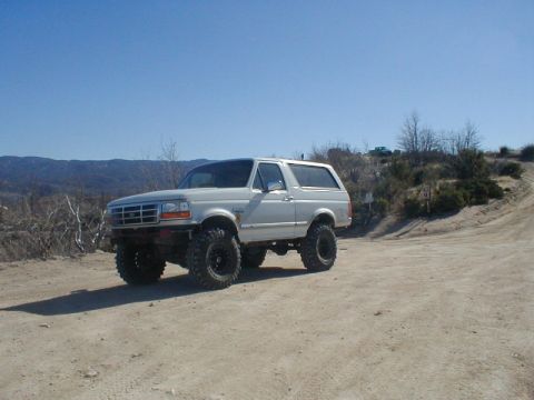 1996 Ford Bronco - A lifted white 1996 Ford Bronco navigates a dusty off-road trail under a clear blue sky.
