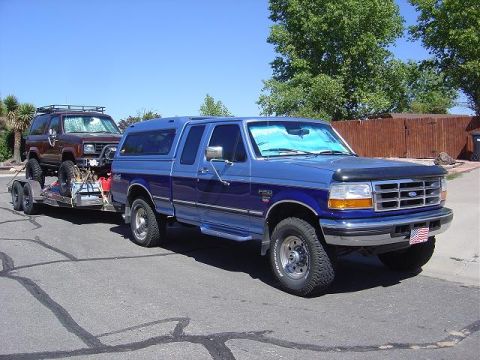 1956 Ford Bronco II - Blue Ford F-250 Super Duty pickup truck tows a brown Ford Bronco on a trailer under a clear blue sky.
