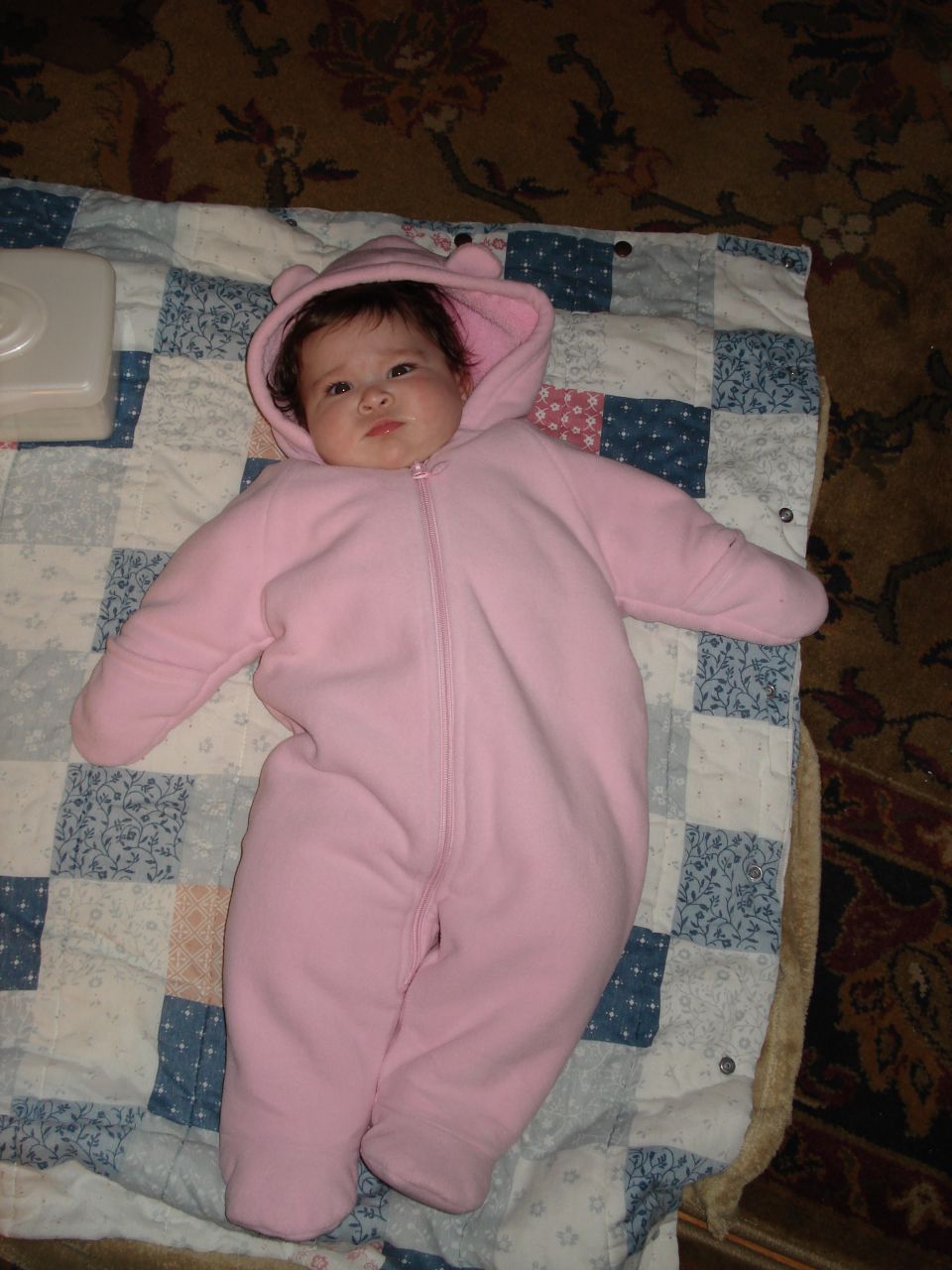 Baby in a pink snowsuit with bear ears on the hood, lying on a quilt