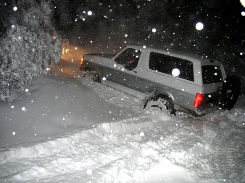 1996 Ford Bronco - A silver two-door Ford Bronco SUV photographed at night during heavy snowfall, viewed from the rear three-quarter angle with its taillights…