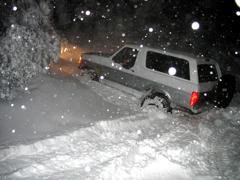 Silver Ford Bronco SUV in deep snow at night with falling snowflakes lit by flash