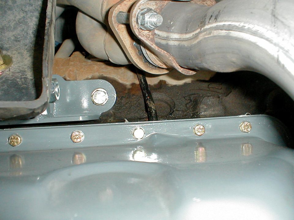 Gray painted oil pan with multiple bolts, connected to an exhaust pipe with a rusty bracket