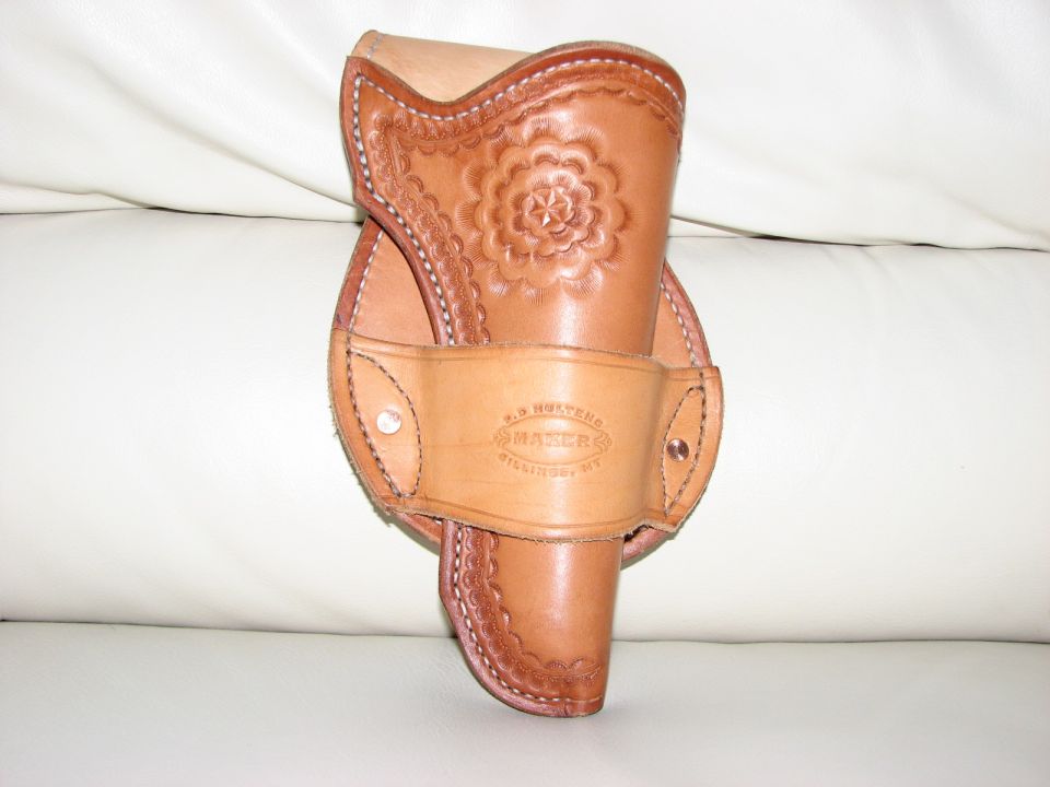 Tan tooled leather Western holster with floral carving and maker stamp, resting on a white leather couch