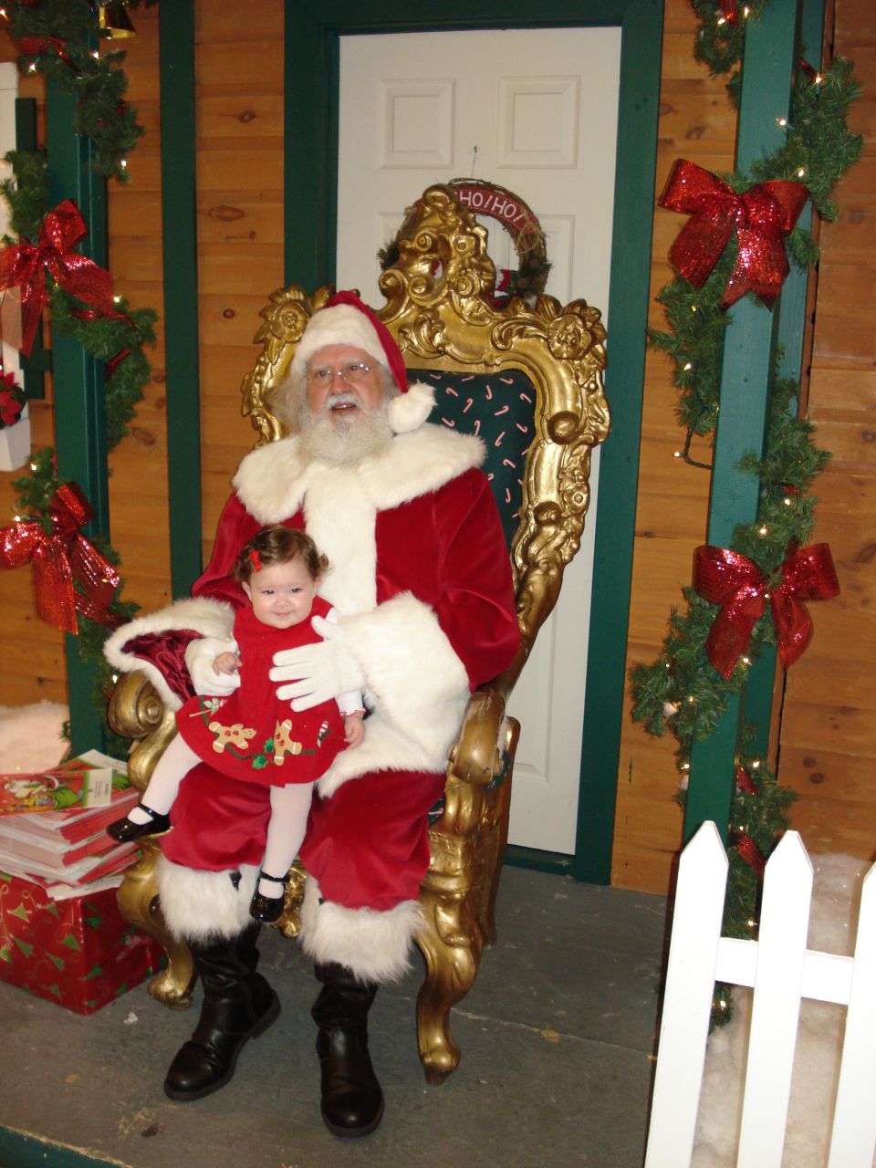 Santa Claus sits in a throne holding a baby dressed in a red holiday outfit. Christmas decorations are visible