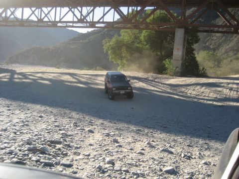 1993 Ford Bronco - An SUV kicks up dust as it drives down a rocky dirt road beneath a large bridge.
