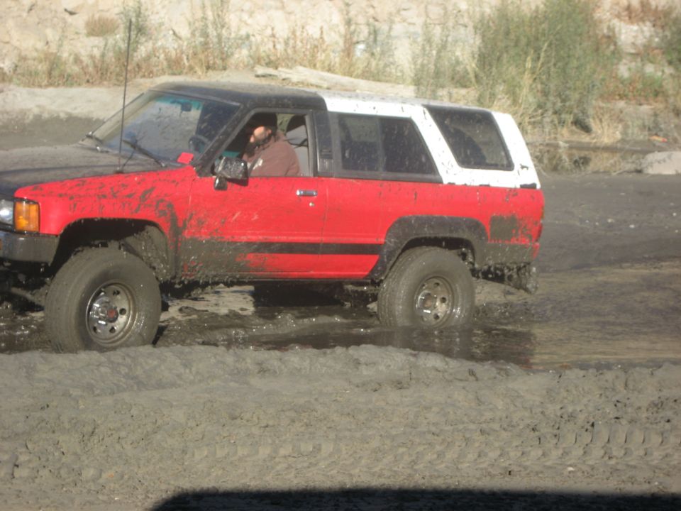Mud-splattered red and black SUV with a white removable top driving through a muddy area