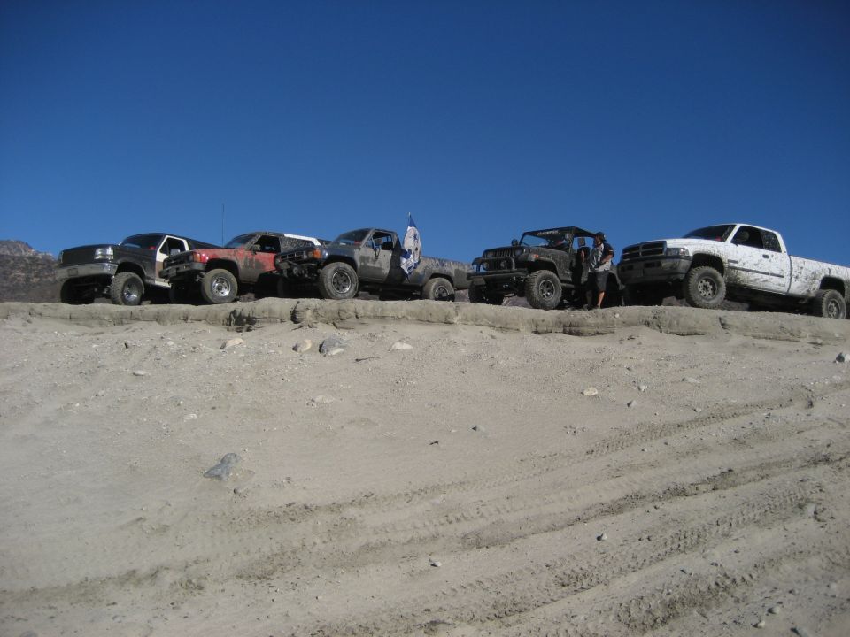 Several off-road trucks and SUVs parked on a sandy ridge under a clear blue sky