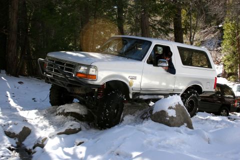 1996 Ford Bronco - A white 1996 Ford Bronco drives through snow and rocks on a wooded trail.