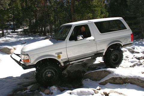 1996 Ford Bronco - A white Ford Bronco is driven over rocks and snow, with the driver visible inside.