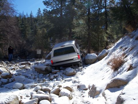 1996 Ford Bronco - A silver SUV navigates a challenging off-road trail covered in snow and rocks.