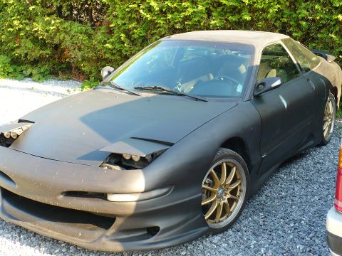 1993 Ford Probe - A matte black 1993 Ford Probe is parked on a gravel surface with trees in the background.