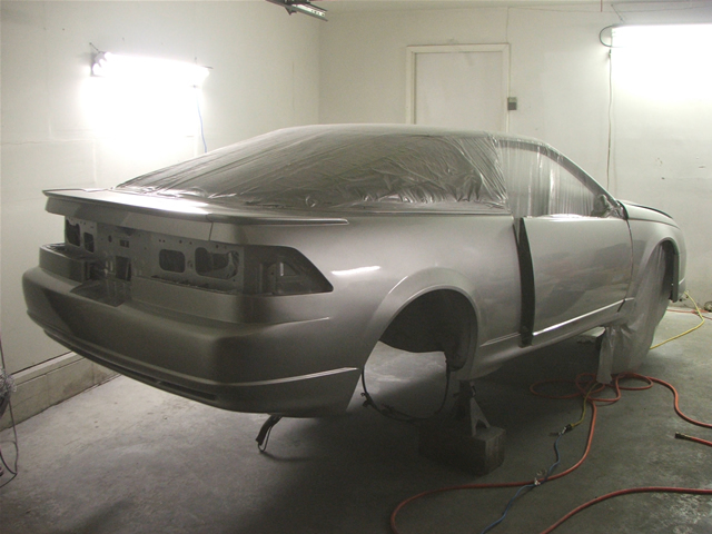 Car body covered in plastic, missing wheels, on stands in a paint booth