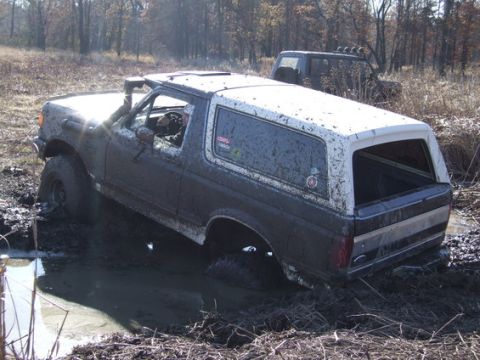 1989 Ford Bronco - Muddy 1989 Ford Bronco is stuck in a water-filled ditch, with another vehicle visible in the background.