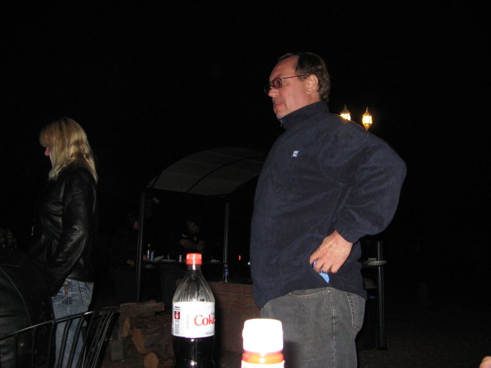 Man in fleece jacket and woman in leather jacket stand at night gathering. Coke bottle in foreground