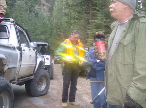 1986 Ford Bronco - People gather around off-road vehicles in a forest setting, with one person holding a large thermos.
