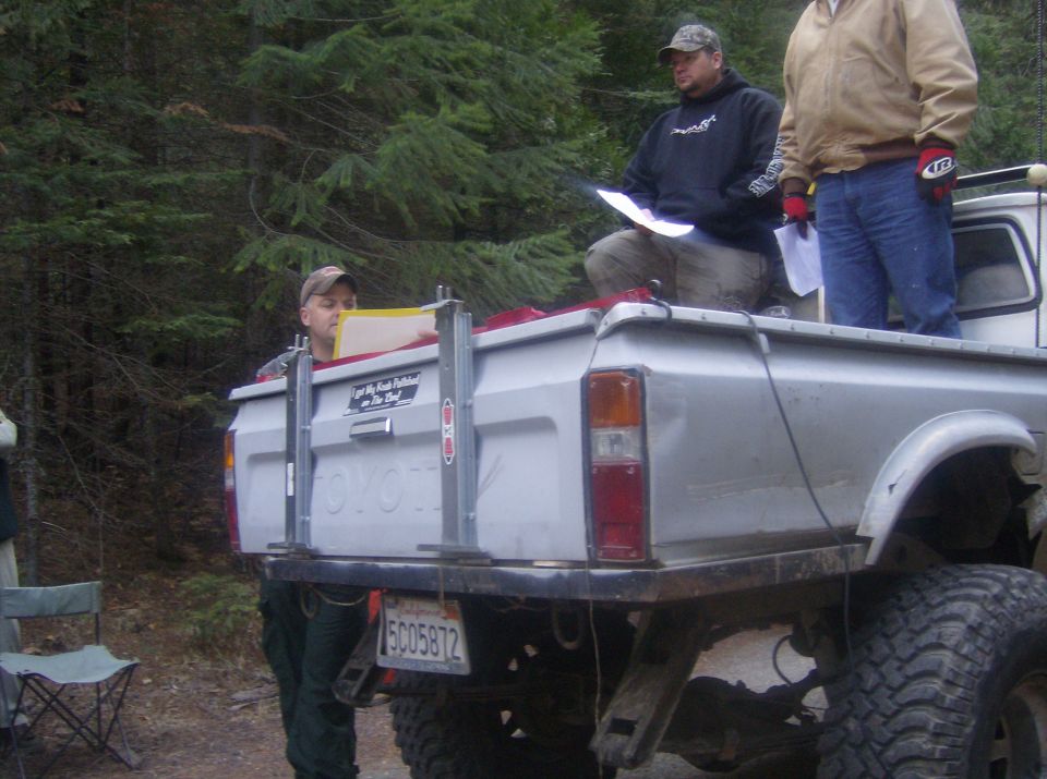 Silver Toyota pickup truck bed with people, large off-road tires, and a license plate reading 5C05872