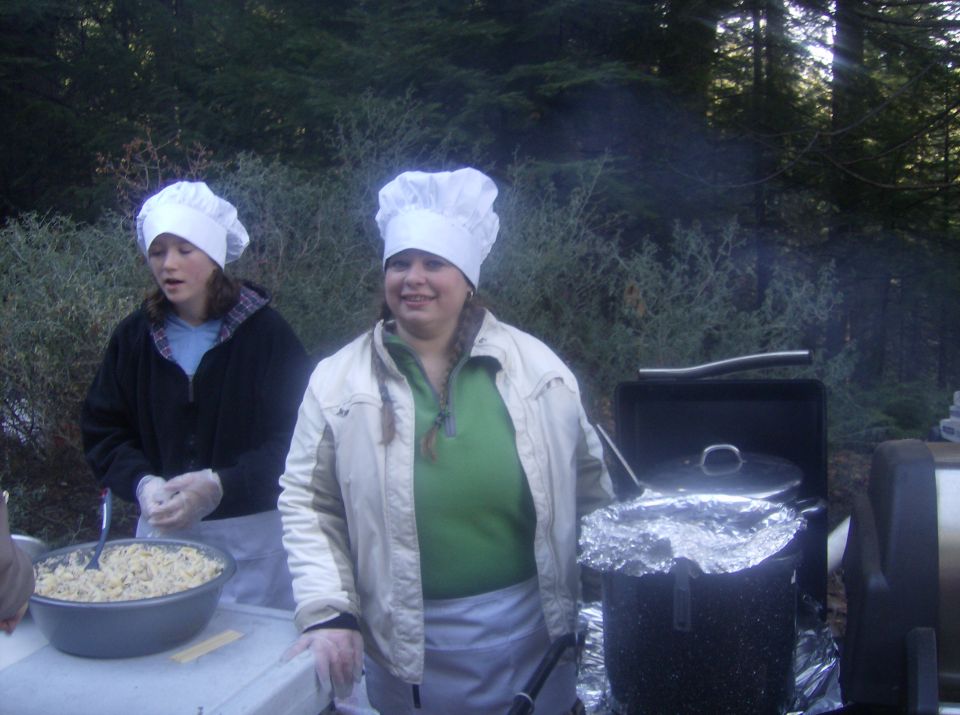 Two people in chef hats prepare food outdoors next to a grill