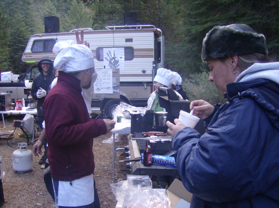 People in chef hats cooking outdoors near an RV, with a propane tank visible