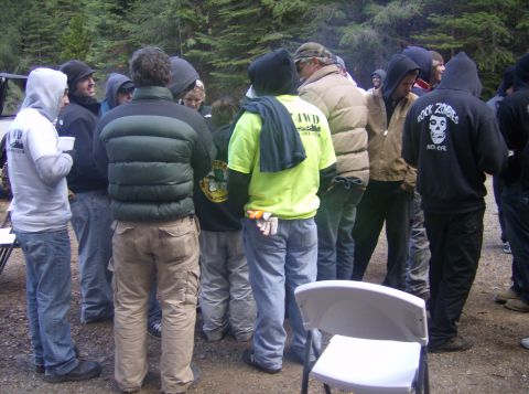 1986 Ford Bronco - People in a wooded area, some wearing hoodies and casual outdoor attire. A white chair is visible in the foreground.
