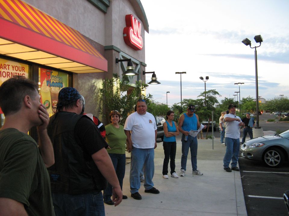 Group of people standing outside a building with a red and yellow awning; a silver car is parked to the right