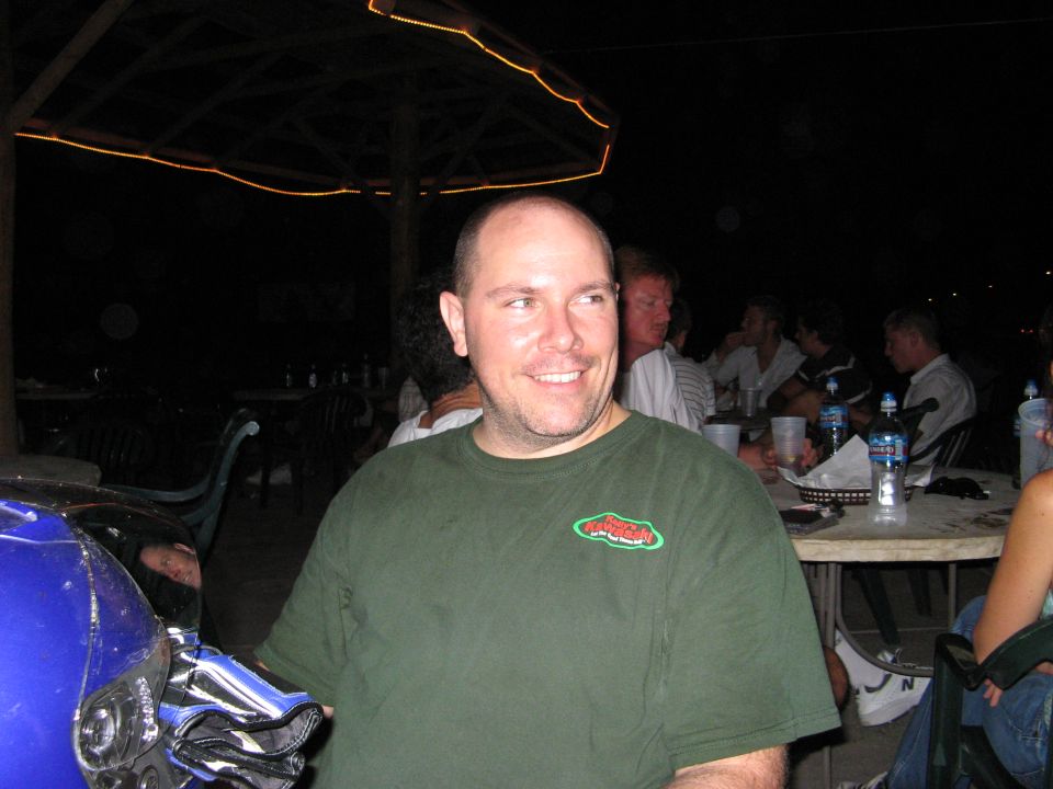 Blue motorcycle fairing in foreground, man in green t-shirt smiling, people at tables in background