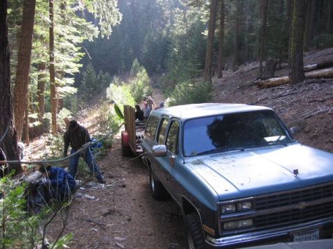 1989 Ford Bronco - A blue SUV tows a trailer down a dirt road in a wooded area, with people working nearby.