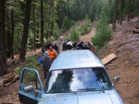 1989 Ford Bronco - A light blue pickup truck is parked on a dirt road in a wooded area, with people gathered around the truck bed.