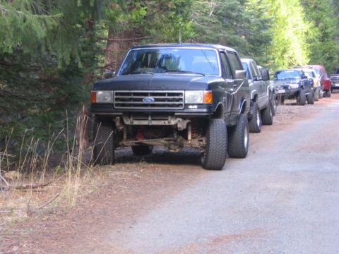 1989 Ford Bronco - Several lifted 4x4 trucks, including a black Ford Bronco, are parked on a dirt road in a wooded area.