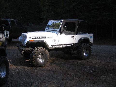 1989 Ford Bronco - A white Jeep Wrangler is parked on a gravel surface with trees in the background. Another vehicle is partially visible to the left.
