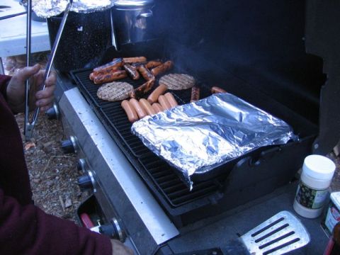 1989 Ford Bronco - Food is being grilled on a barbecue, including hot dogs and hamburgers, with a dish covered in aluminum foil.
