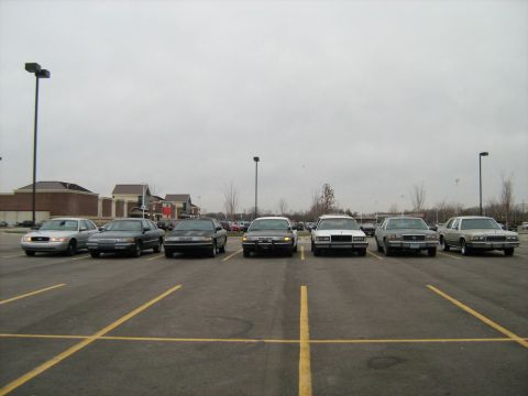 1988 Mercury Grand Marquis - A line of six sedans, including a 1988 Mercury Grand Marquis, are parked in a lot under an overcast sky.