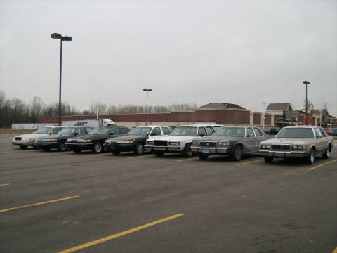 1988 Mercury Grand Marquis - Several 1988 Mercury Grand Marquis sedans are lined up in a parking lot under an overcast sky.