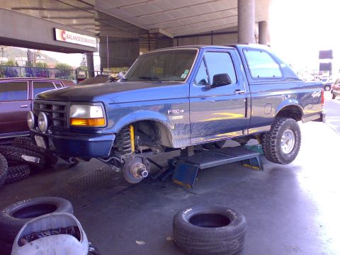 1995 Ford F-150 - Blue 1990s Ford F-150 with an F-150 V8 fender badge and 4x4 bedside decal, jacked up on a tire-shop alignment rack with its front wheel rem…