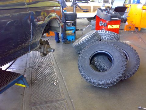 1995 Ford F-150 - Tire shop scene with a dark green Ford F-150 (V8 and F-150 fender badges) raised on a jack stand, front wheel removed to expose the hub, ro…