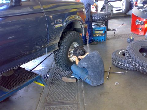 1995 Ford F-150 - A dark-colored Ford F-150 pickup with a V8 badge is raised on a shop lift while a person crouches to work on the front wheel fitted with an…