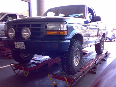 1995 Ford F-150 - Green Ford F-150 4x4 pickup raised on a shop alignment rack, fitted with off-road lights and mud-terrain tires.