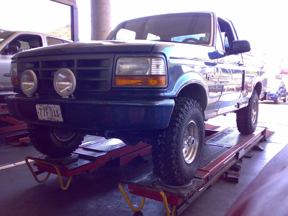 Green Ford F-150 4x4 pickup on shop alignment rack with off-road lights and mud tires