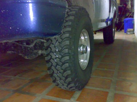 1995 Ford F-150 - Low-angle close-up of a dark blue Ford pickup's front wheel fitted with an aggressive mud-terrain tire on a polished aluminum wheel, shot o…