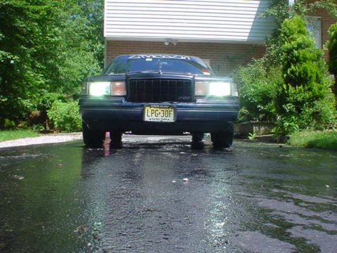 1994 Lincoln Town Car - Front view of a dark sedan with its headlights on, parked on a wet surface in front of a building.
