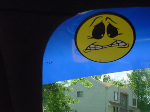 1994 Lincoln Town Car - A worried-looking smiley face sticker adorns the blue visor of a car, with a house and trees visible through the windshield.