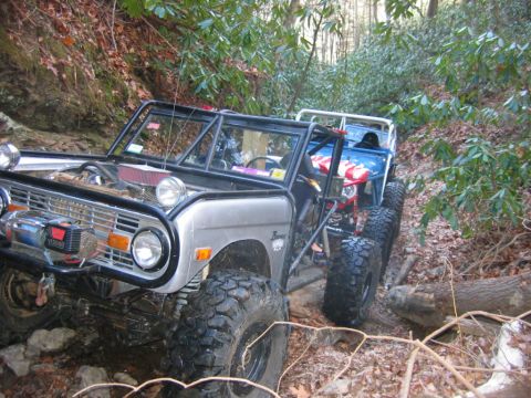 1992 Ford Mustang - Two modified off-road vehicles navigate a challenging wooded trail with steep inclines and fallen logs.