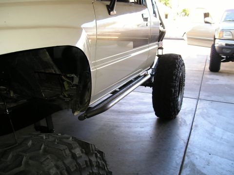 1986 Toyota Hilux - Low side view of a lifted white pickup in a driveway with bare-steel tube rock sliders, black exocage bars at the cab, and the bed removed…