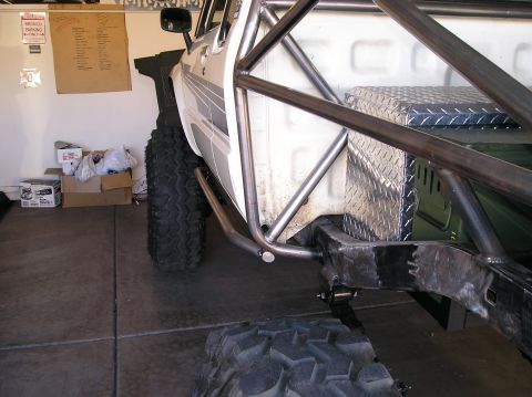 1986 Toyota Hilux - Bare-steel exo/roll cage tubing being fabricated along the side of a white Toyota pickup in a garage, tying into the bare frame rails behin…