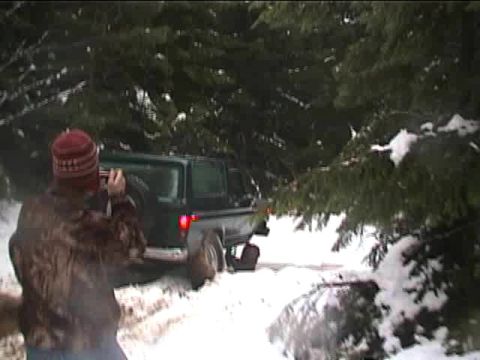 1979 Ford Bronco - A person films a green SUV navigating a snow-covered trail surrounded by dense pine trees.